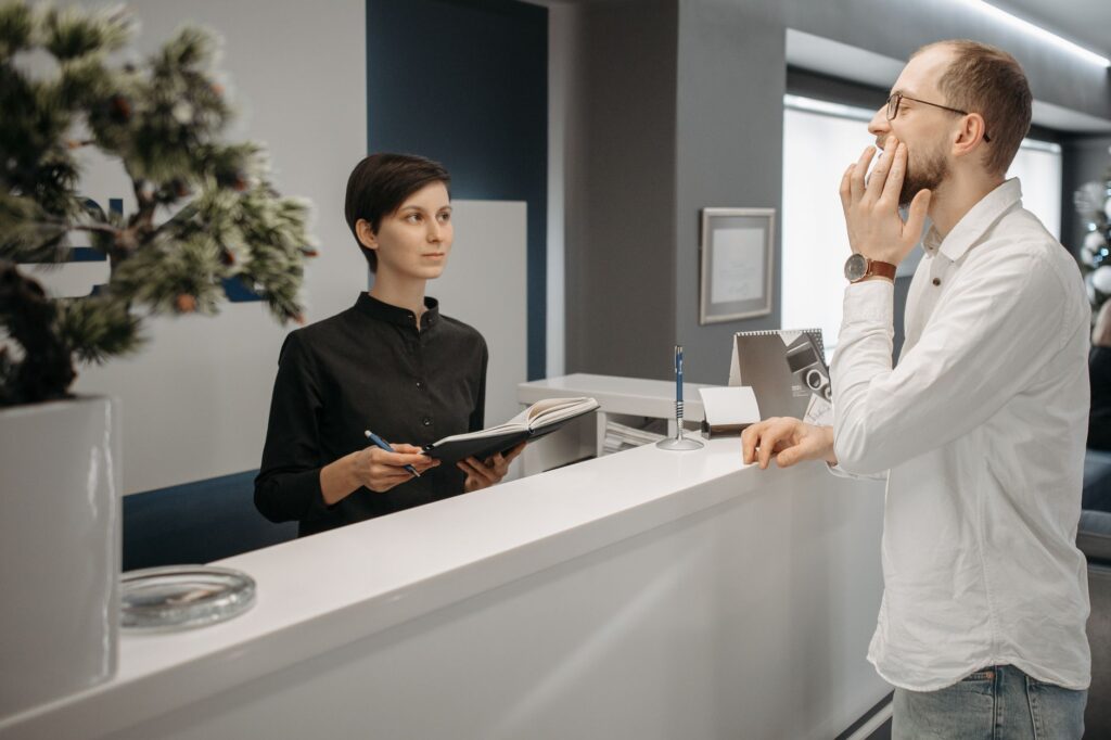 Dental office receptionist with an appointment book helping a patient who has a toothache, the point where a Google Ads lead becomes a booked visit
